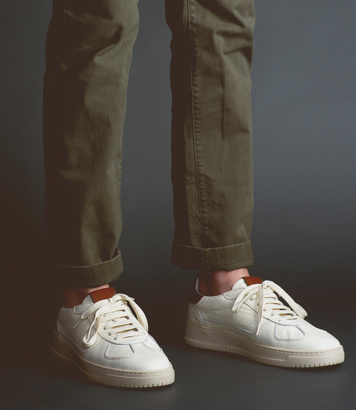 Wearing olive green pants and classic men’s footwear—the sleek "North" white sneakers by Bed|Stü—a person stands against a dark background.