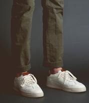 Wearing olive green pants and classic men’s footwear—the sleek "North" white sneakers by Bed|Stü—a person stands against a dark background.