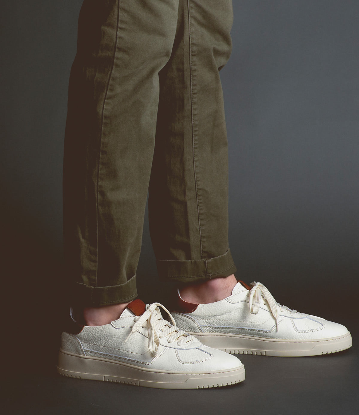 A person wearing rolled-up olive green pants and classic men's footwear—the Bed|Stü North Italian leather sneakers in crisp white—stands on a dark floor against a plain gray background.