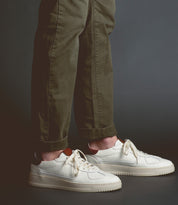 A person wearing rolled-up olive green pants and classic men's footwear—the Bed|Stü North Italian leather sneakers in crisp white—stands on a dark floor against a plain gray background.
