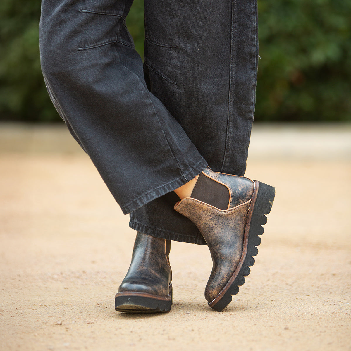 Someone wearing Bed|Stü's Novel II boots—Chelsea-style with a worn leather upper—stands outdoors on a dirt pathway, one foot crossed over the other, paired with loose, dark pants.