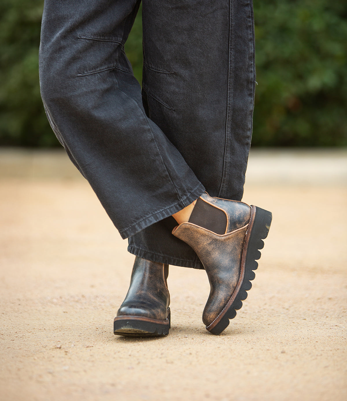Someone wearing Bed|Stü's Novel II boots—Chelsea-style with a worn leather upper—stands outdoors on a dirt pathway, one foot crossed over the other, paired with loose, dark pants.