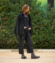 A person is standing outdoors on a dirt surface, wearing black overalls, a black fringed jacket, a purple turtleneck, and the Bed|Stü Novel II dark Chelsea boots with a platform sole and leather upper, with greenery in the background.