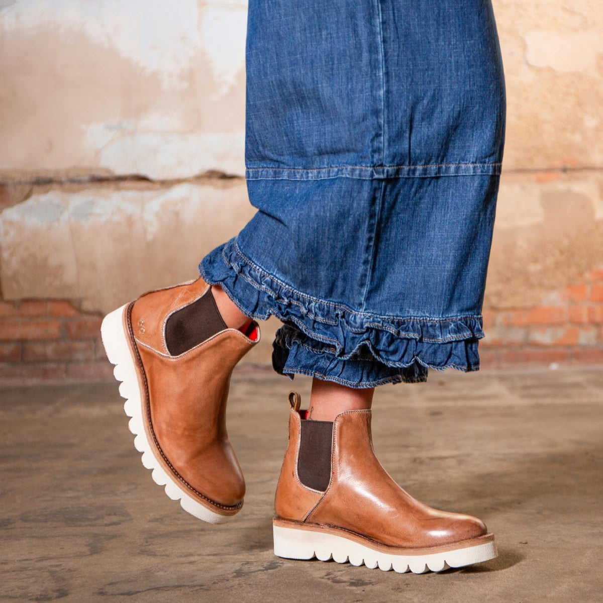 Person wearing frayed-hem blue jeans and Bed|Stü Novel II tan Chelsea boots with a leather upper and white soles, standing on a concrete floor with a brick and plaster wall in the background.