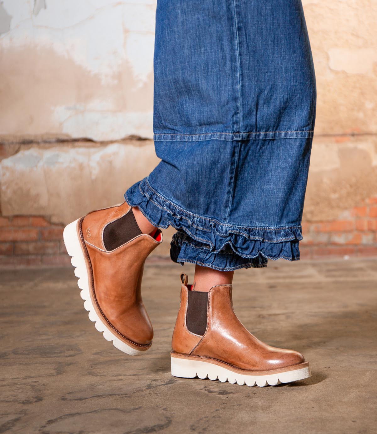 Person wearing frayed-hem blue jeans and Bed|Stü Novel II tan Chelsea boots with a leather upper and white soles, standing on a concrete floor with a brick and plaster wall in the background.