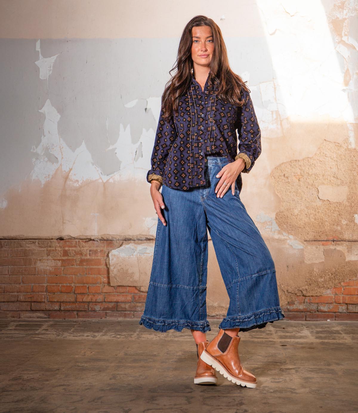 A woman stands indoors against a worn, textured wall, wearing a patterned blouse, wide-leg denim pants, and Bed|Stü’s Novel II brown Chelsea boots with a stylish platform sole.