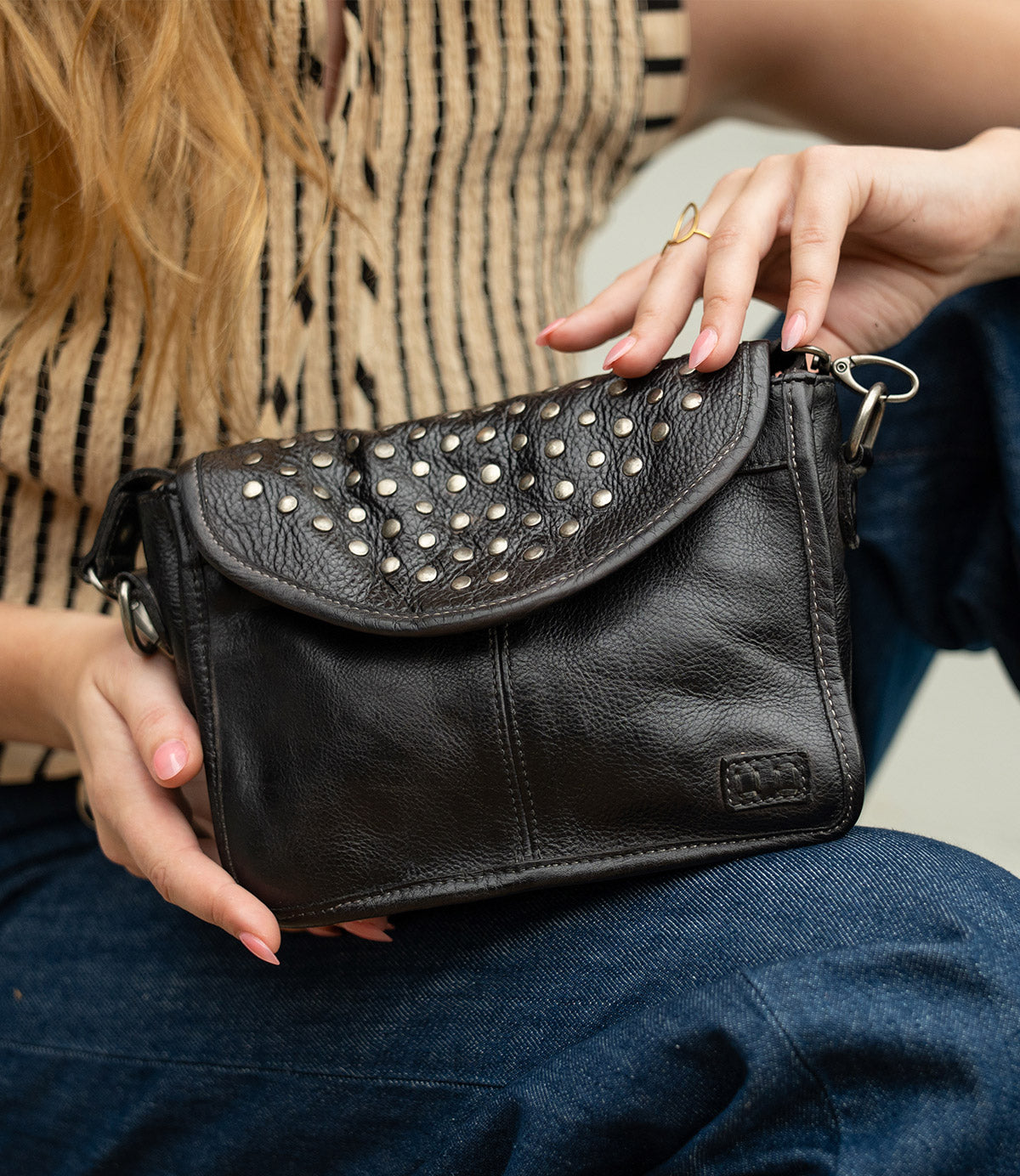 Wearing a striped sleeveless top and jeans, the person holds the Bed|Stü Occurrence—a compact crossbody bag featuring metal stud accents and a flap closure.