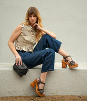 A woman sits on a ledge wearing a sleeveless striped top, wide-leg jeans, and wooden platform sandals, holding the Bed|Stü Occurrence—a black studded bag that adds an edgy and versatile touch to her look.