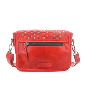 The Bed|Stü Occurrence is a red leather compact crossbody bag featuring metal studs on the flap, a front zipper pocket, and a detachable shoulder strap, shown against a white background.
