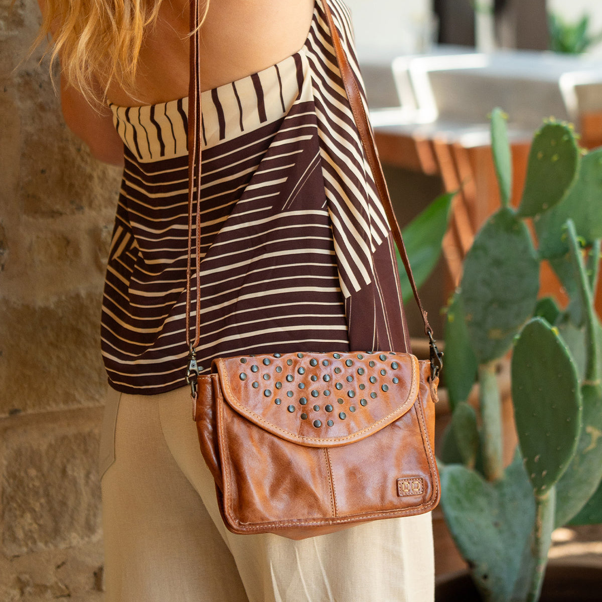 A person wearing a striped top and beige pants stands by a stone wall, carrying the Bed|Stü Occurrence crossbody bag—a compact accessory crafted from brown leather with studded details that perfectly complements the outfit. A cactus is visible in the background.