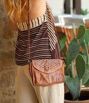 A person wearing a striped top and beige pants stands by a stone wall, carrying the Bed|Stü Occurrence crossbody bag—a compact accessory crafted from brown leather with studded details that perfectly complements the outfit. A cactus is visible in the background.