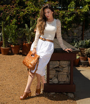 A woman stands outdoors by a stone table, holding the Bed|Stü Orchid handwoven leather bag. She wears a cream lace top and white skirt, while potted plants and lush greenery in the background lend a modern timeless feel to the tranquil setting.