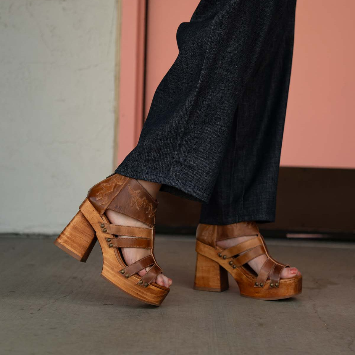 A person is wearing dark denim pants and the Orphyx heels by Bed|Stü, which feature a sculpted wood platform, layered brown leather straps, and an open-toe silhouette, standing on a concrete floor near a pale pink door.