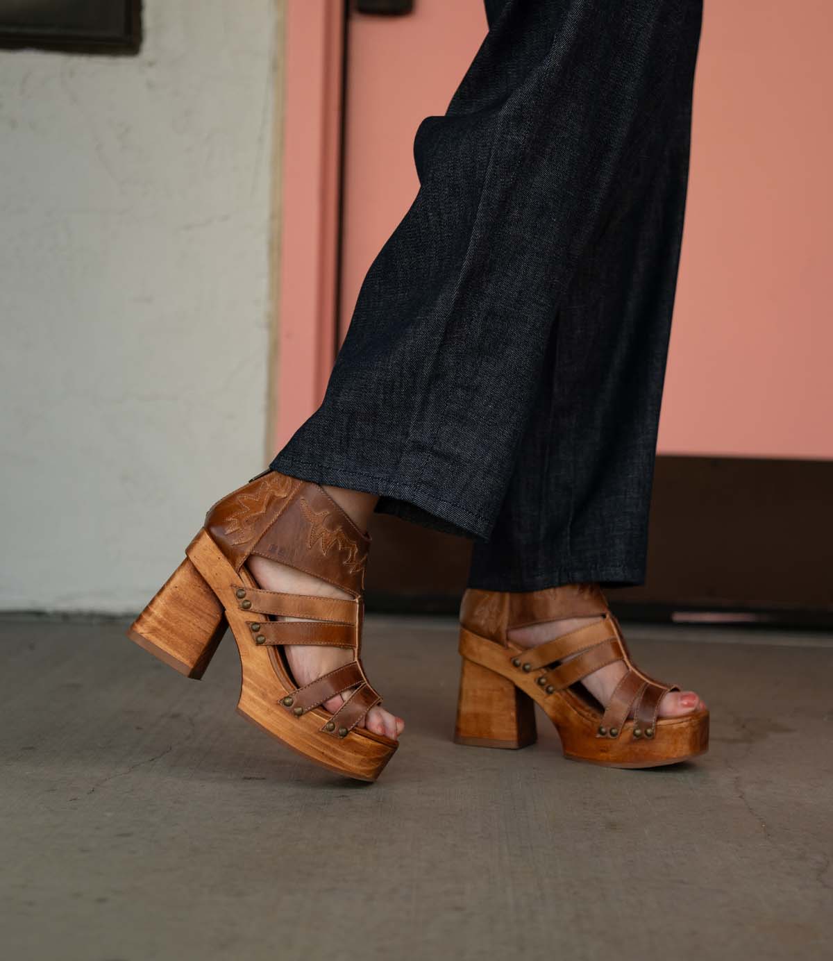A person is wearing dark denim pants and the Orphyx heels by Bed|Stü, which feature a sculpted wood platform, layered brown leather straps, and an open-toe silhouette, standing on a concrete floor near a pale pink door.
