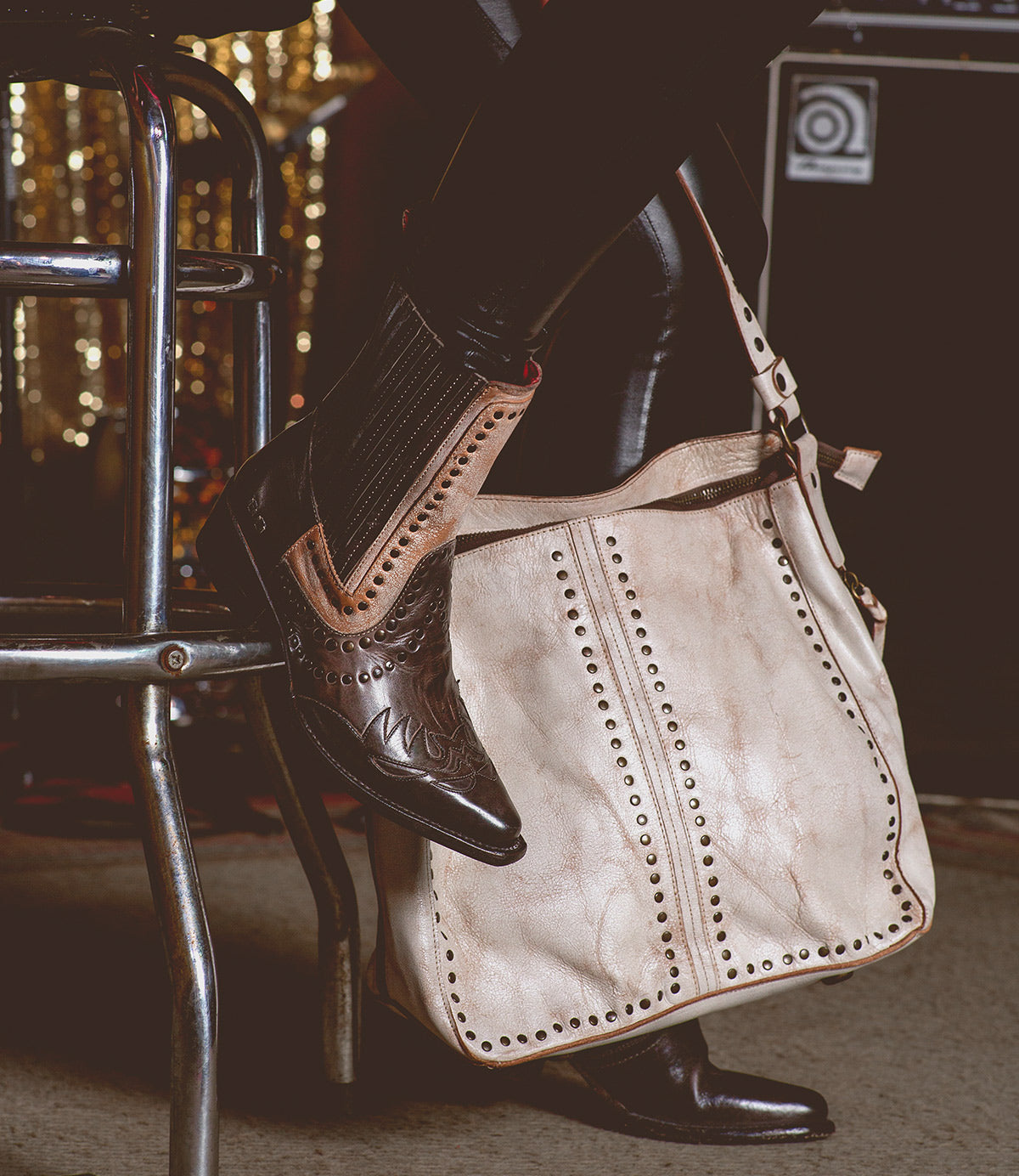 Wearing brown Bed|Stü cowboy boots and black pants, the person sits on a metal stool with the large beige Packet LTC leather tote by their side.