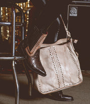 Wearing brown Bed|Stü cowboy boots and black pants, the person sits on a metal stool with the large beige Packet LTC leather tote by their side.