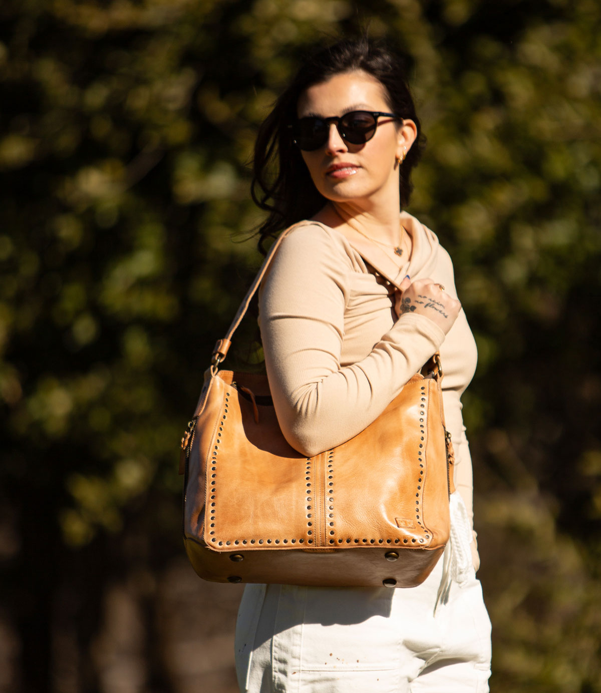 A woman wearing sunglasses and a beige top stands outdoors with blurred greenery in the background, carrying the Bed|Stü Packet LTC—a large tan leather tote—over her shoulder.