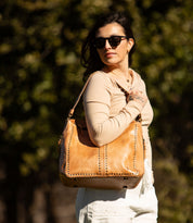 A woman wearing sunglasses and a beige top stands outdoors with blurred greenery in the background, carrying the Bed|Stü Packet LTC—a large tan leather tote—over her shoulder.