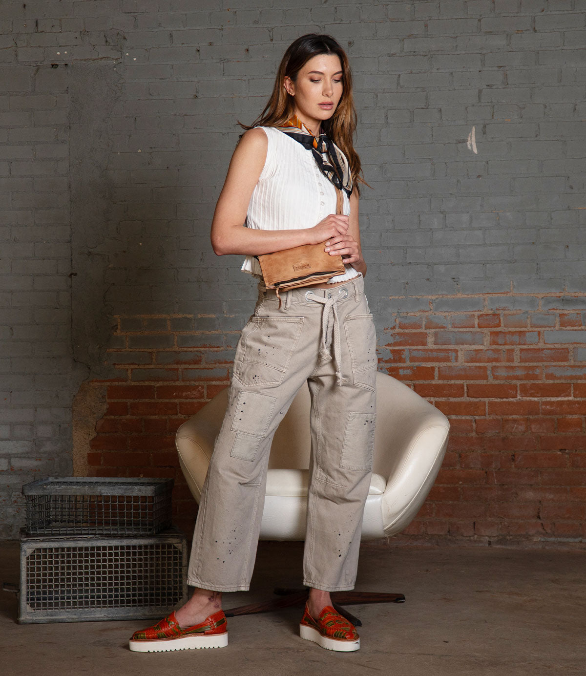 A woman stands indoors near a white chair and stacked wire baskets against a brick and gray wall, dressed in a white sleeveless top, beige cargo pants, red patterned shoes, and a scarf. Effortlessly draped across her shoulder by its crossbody strap is the Bed|Stü Parle, a soft leather bag featuring zippered compartments.