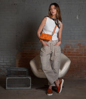 A woman stands in front of a gray brick wall, wearing a white sleeveless top, beige cargo pants, orange shoes, and carrying the Bed|Stü Parle orange soft leather bag with zippered compartments and a crossbody strap, with one hand in her pocket.