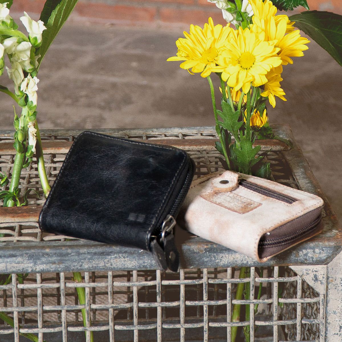 The Peek by Bed|Stü, a black zippered wallet crafted from premium leather, and a beige card holder featuring accordion-style slots rest on a metal crate, with yellow flowers and greenery in the background.