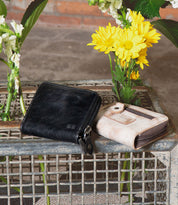 The Peek by Bed|Stü, a black zippered wallet crafted from premium leather, and a beige card holder featuring accordion-style slots rest on a metal crate, with yellow flowers and greenery in the background.