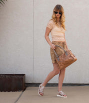 A woman in sunglasses, dressed in a lace top, khaki shorts, and white platform sandals with leather straps, carries the Bed|Stü Perfect tan handbag as she walks on a sidewalk in front of a light-colored textured wall.