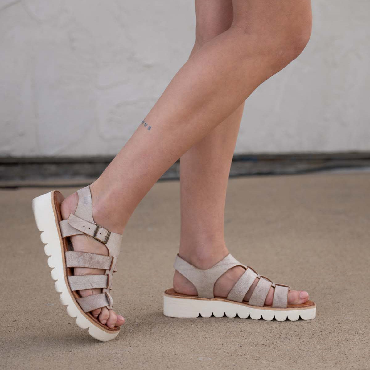 A person wearing Bed|Stü Perfect light beige leather platform sandals with thick white soles stands on a concrete surface. Only their legs and feet are visible, highlighting the stylish leather straps of these fashionable fisherman sandals.