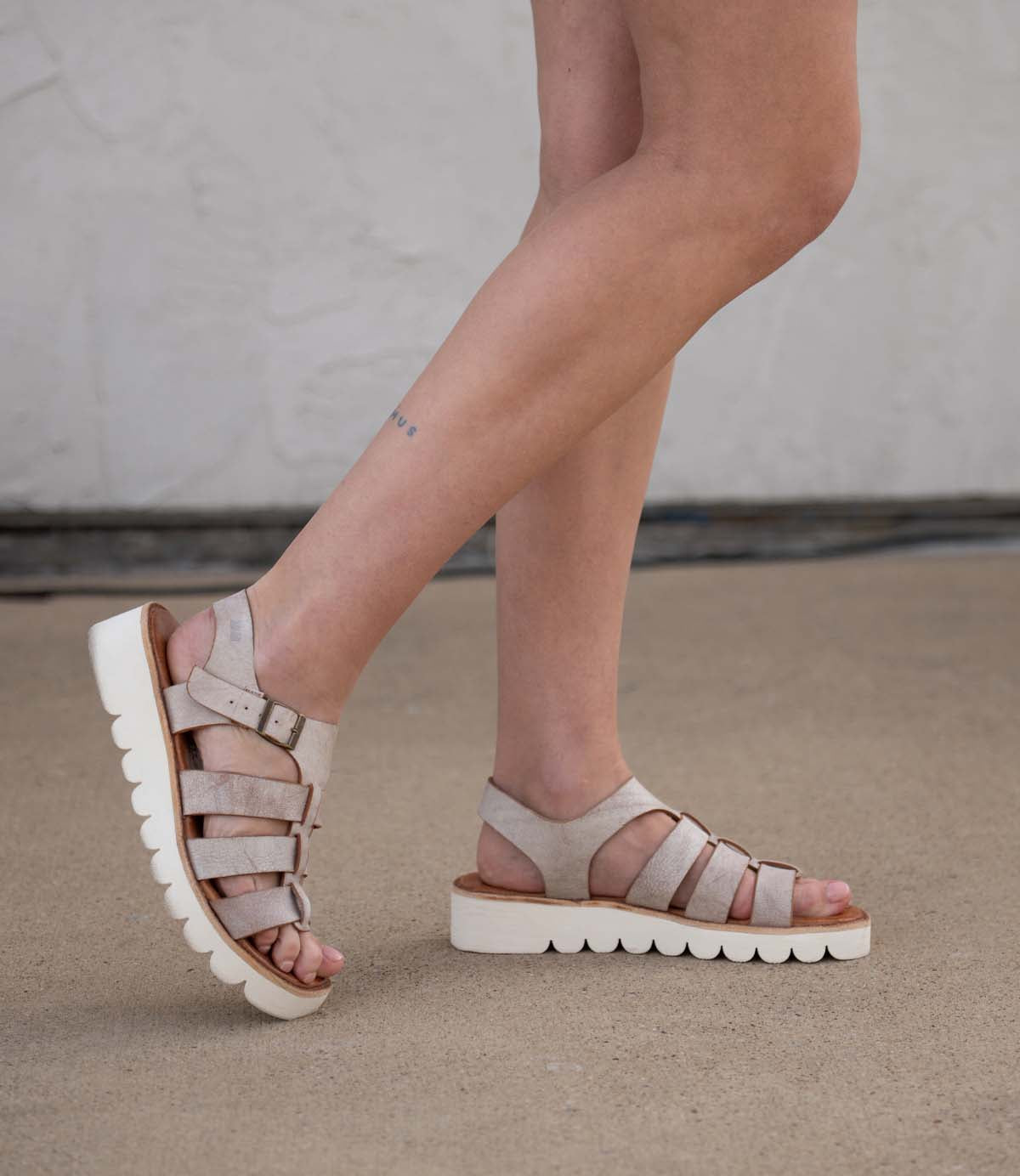 A person wearing Bed|Stü Perfect light beige leather platform sandals with thick white soles stands on a concrete surface. Only their legs and feet are visible, highlighting the stylish leather straps of these fashionable fisherman sandals.