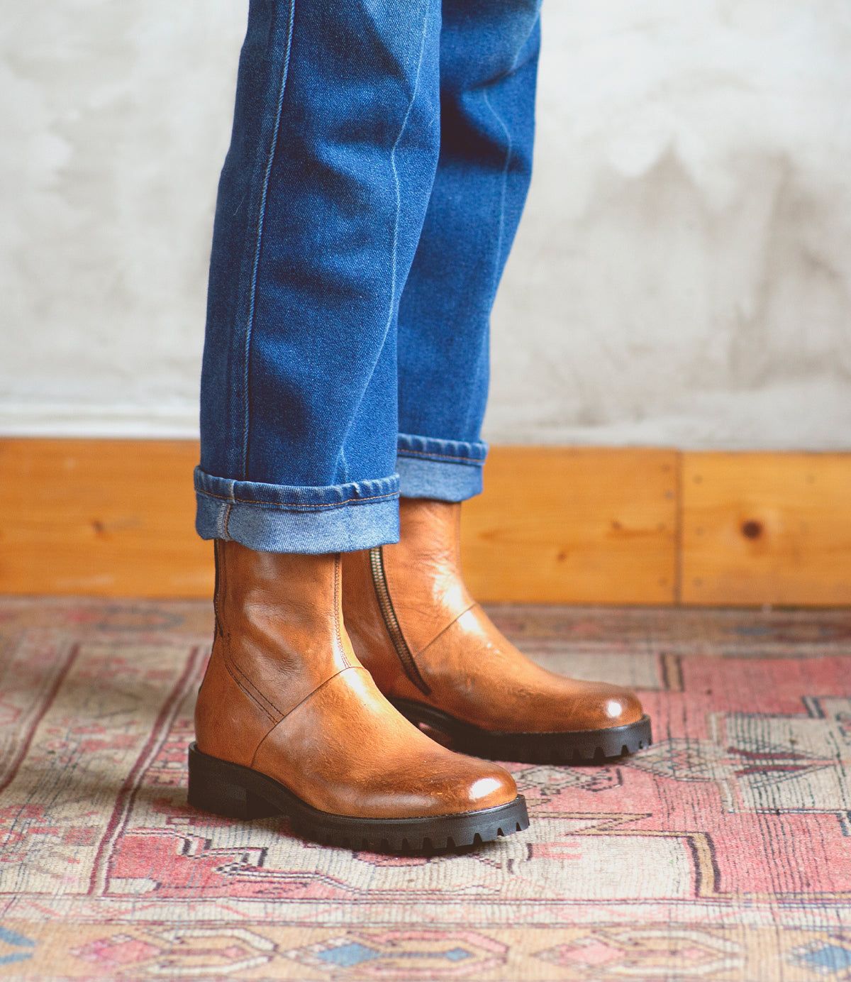 A person wearing blue jeans and the Ploy ankle boots by Bed|Stü—crafted from tan Italian leather with square toes and subtle dual side zippers for both style and convenience—stands on a patterned rug, with a wooden baseboard and textured wall in the background.