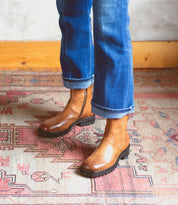 Wearing the Bed|Stü Ploy boots—stylish brown Italian leather ankle boots with dual side zippers—a person in cuffed blue jeans stands on a patterned rug, framed by a wooden baseboard in the background.