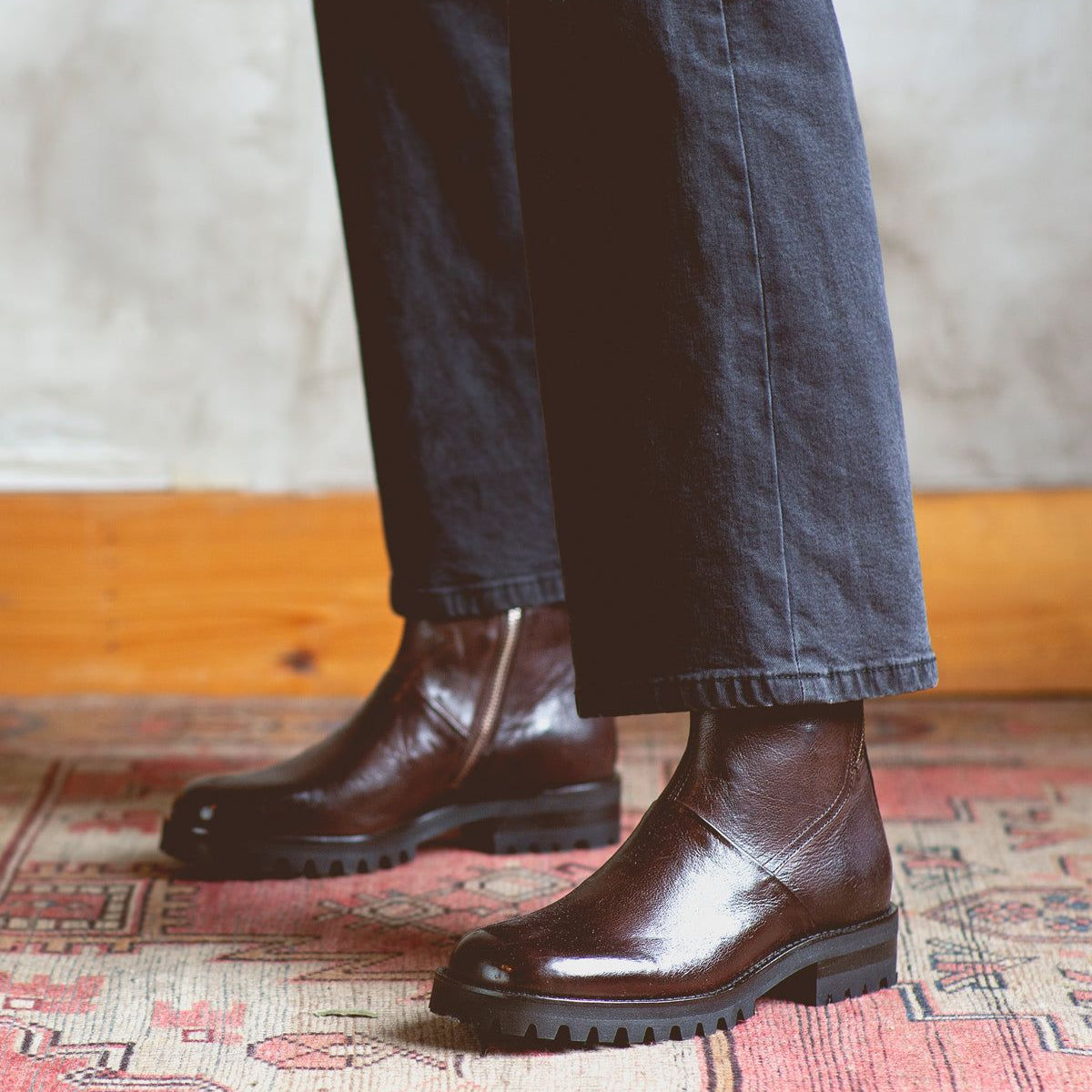 A person wearing the Bed|Stü Ploy dark brown Italian leather boots with thick soles and black pants stands on a patterned rug, with a light-colored wall in the background.