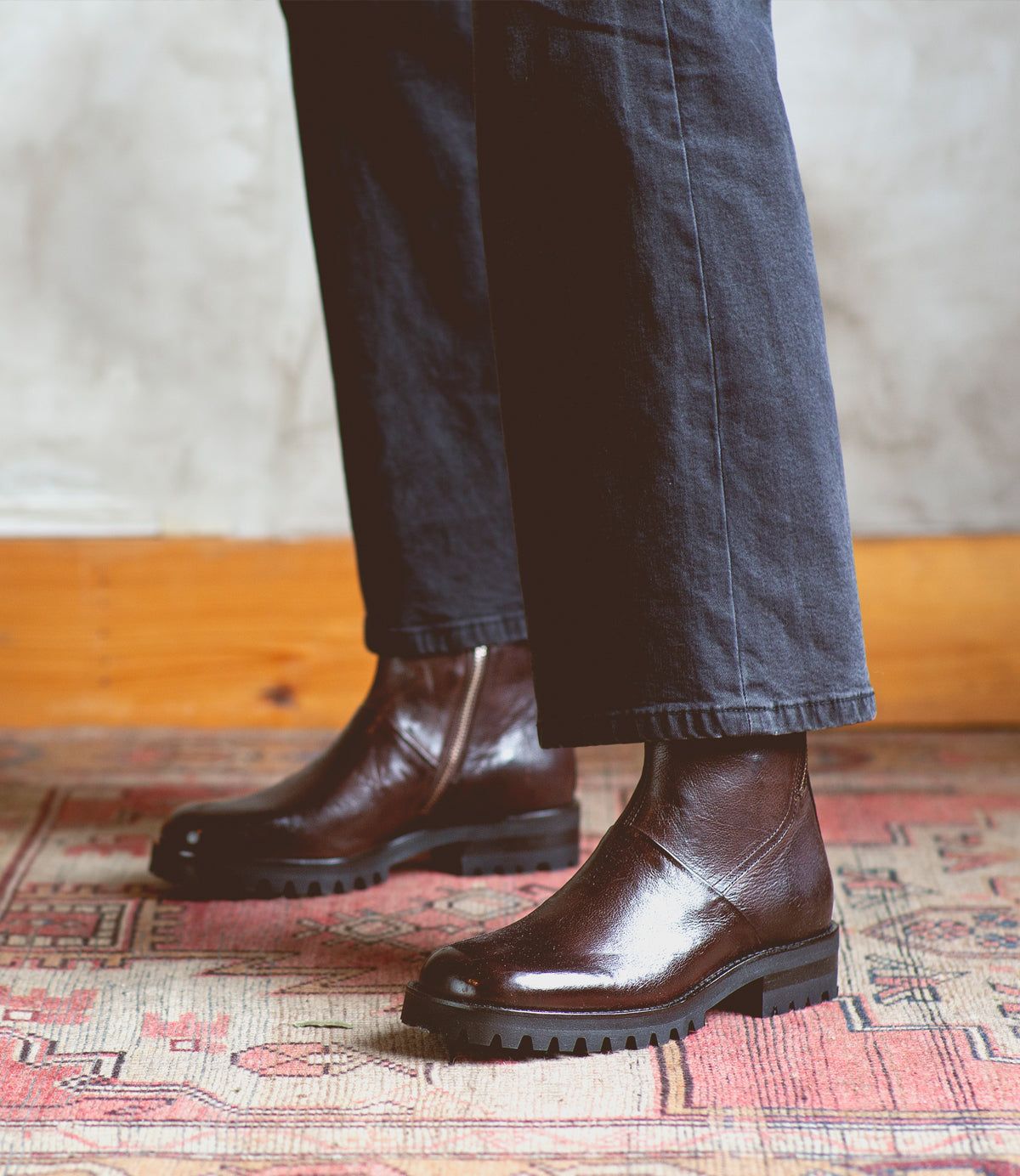 A person wearing the Bed|Stü Ploy dark brown Italian leather boots with thick soles and black pants stands on a patterned rug, with a light-colored wall in the background.