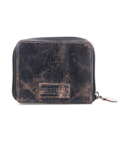 A gently worn black compact leather wallet by Bed|Stü, the Plus model, with visible creases and a textured finish. It features a stitched rectangular patch on the front, a zippered coin pocket, and a zipper pull on the right side.