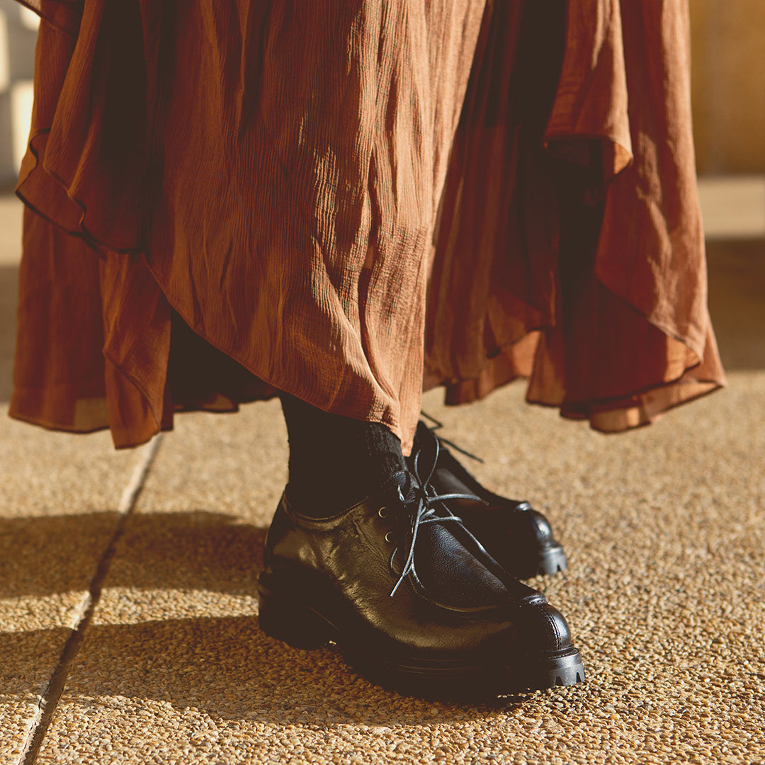 A person stands on textured ground wearing a long brown skirt, black socks, and Bed|Stü Portare boots styled as vintage-inspired moc-toe lace-up boots.
