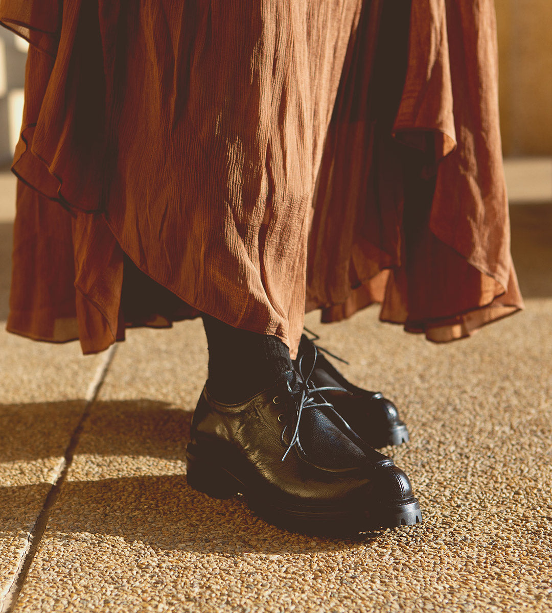 A person stands on textured ground wearing a long brown skirt, black socks, and Bed|Stü Portare boots styled as vintage-inspired moc-toe lace-up boots.