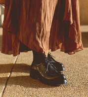 A person stands on textured ground wearing a long brown skirt, black socks, and Bed|Stü Portare boots styled as vintage-inspired moc-toe lace-up boots.
