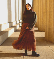 A woman in a green sweater and brown skirt stands in sunlight beside tall stone columns, looking at the camera, her Bed|Stü Portare boots adding a timeless, vintage-inspired touch to the ensemble.