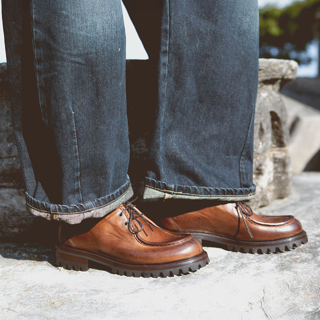 A person wearing dark jeans and a pair of Bed|Stü Portare vintage-inspired moc-toe lace-up boots with thick black soles stands on a stone surface outdoors.