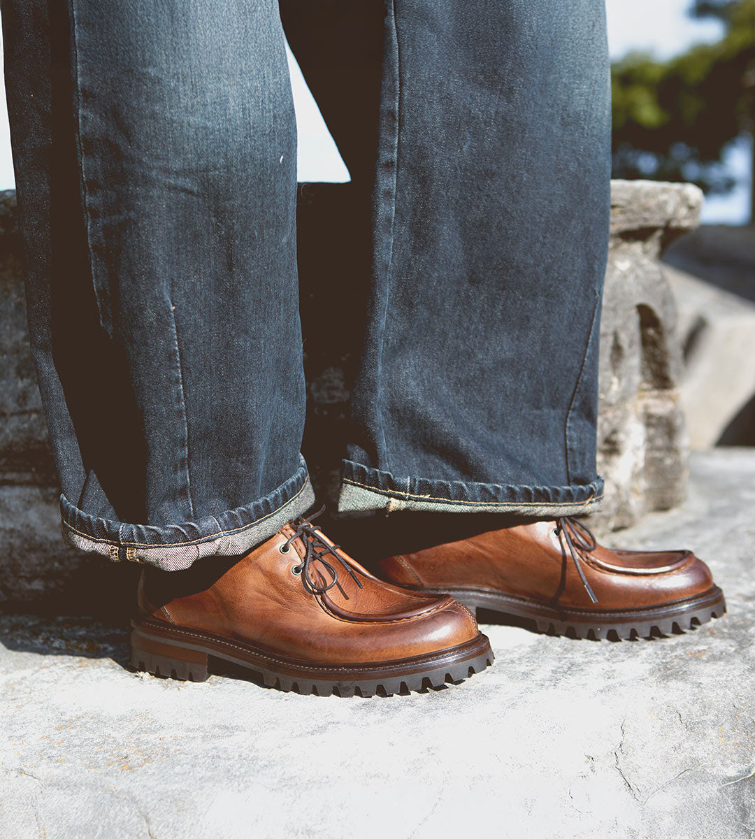 A person wearing dark jeans and a pair of Bed|Stü Portare vintage-inspired moc-toe lace-up boots with thick black soles stands on a stone surface outdoors.