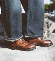 A person wearing dark jeans and a pair of Bed|Stü Portare vintage-inspired moc-toe lace-up boots with thick black soles stands on a stone surface outdoors.