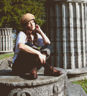 Wearing the Bed|Stü Portare boots, a person in a tan beret sits on a stone structure near large stone columns outdoors, gently touching their face.