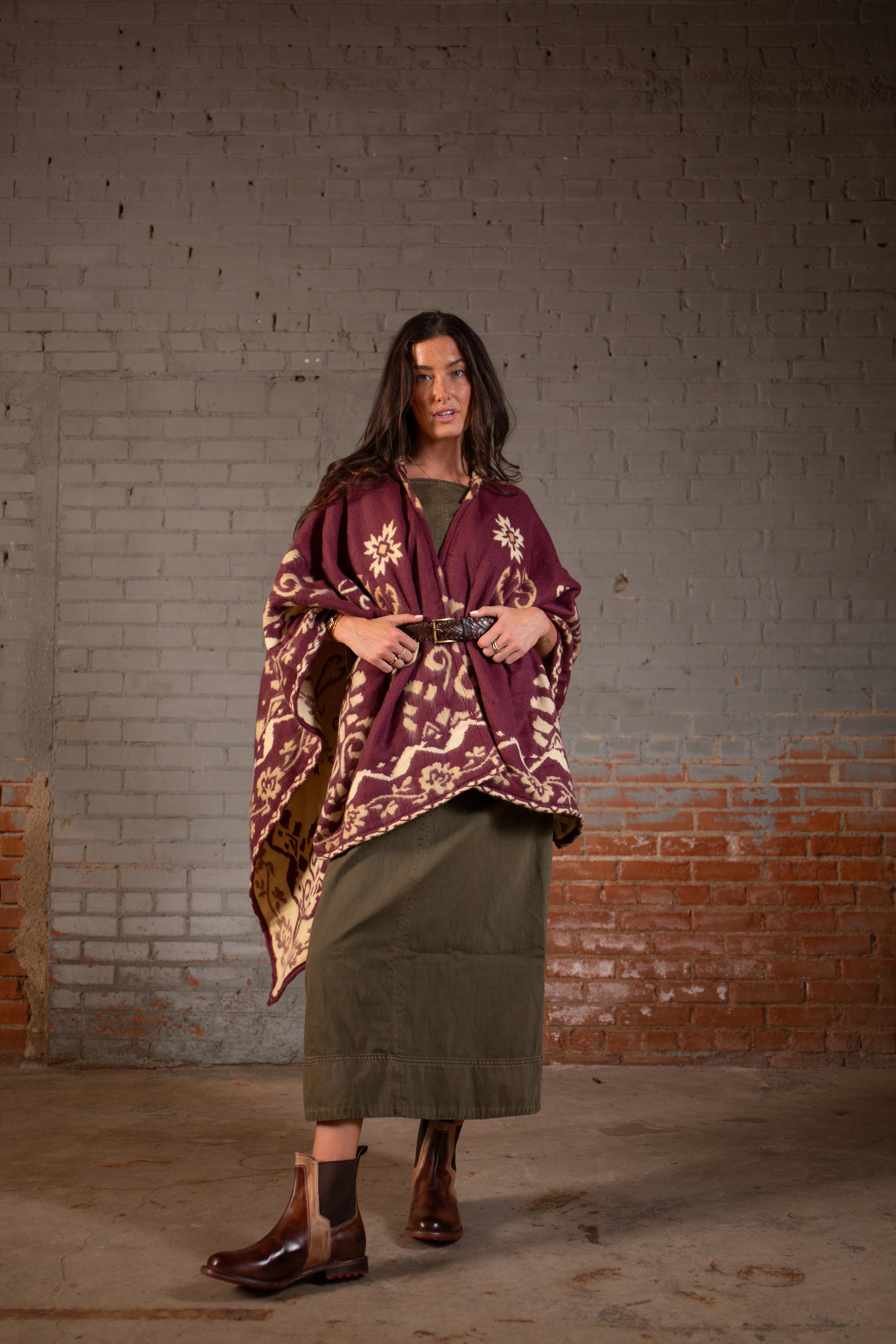 A woman stands indoors posed against a brick and gray painted wall, wearing a patterned burgundy poncho, green skirt, and the Bed|Stü Proem handwoven leather belt with a vintage metal buckle, paired with brown boots.