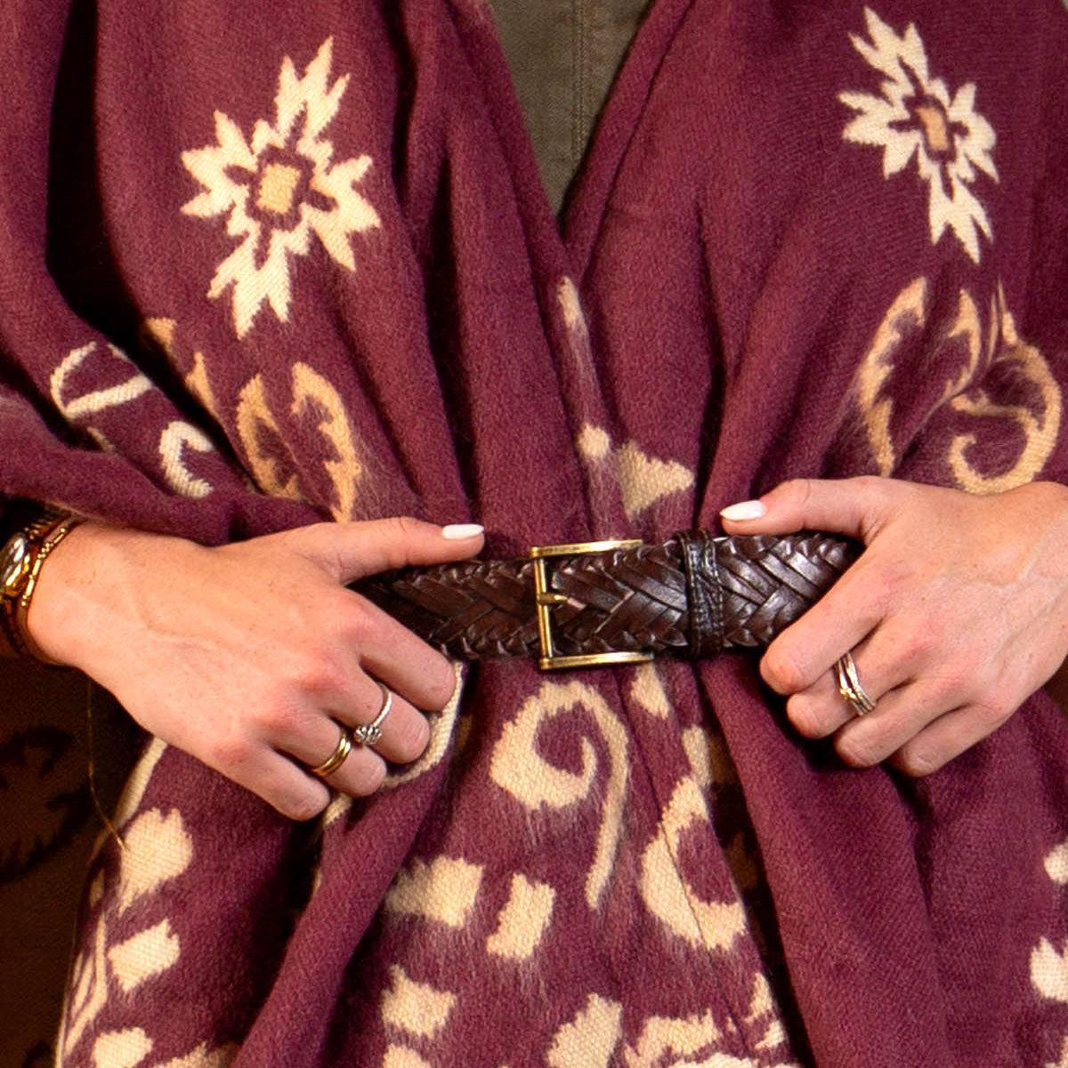 A person wearing a maroon and cream patterned shawl, cinched at the waist with the Bed|Stü Proem handwoven leather belt featuring artisan detailing; their hands display pale nail polish and rings.