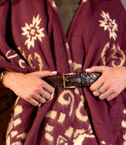 A person wearing a maroon and cream patterned shawl, cinched at the waist with the Bed|Stü Proem handwoven leather belt featuring artisan detailing; their hands display pale nail polish and rings.