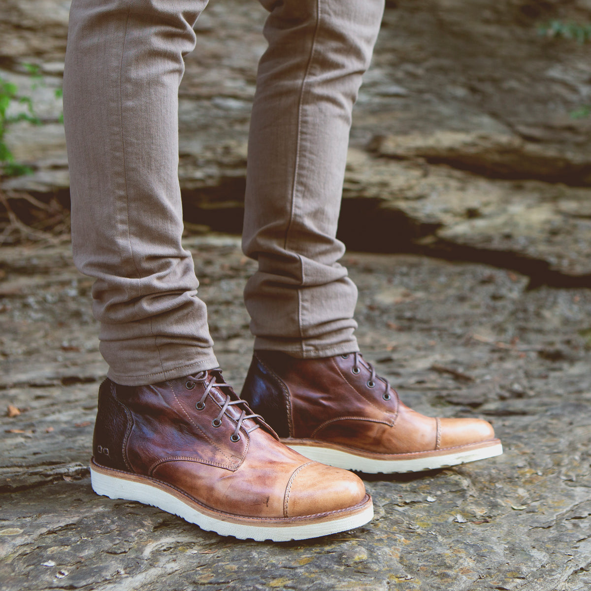 A person wearing Bed|Stü Protege Light brown leather boots with a lightweight, shock-absorbing outsole and light brown pants stands on a rocky outdoor surface.