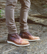 A person wearing Bed|Stü Protege Light brown leather boots with a lightweight, shock-absorbing outsole and light brown pants stands on a rocky outdoor surface.