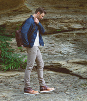 A man wearing a denim jacket, beige pants, a backpack, and Bed|Stü Protege Light boots—which feature Goodyear welt construction and a lightweight, shock-absorbing outsole—stands on rocky ground near a stone wall with green plants, creating an ideal look for everyday wear.