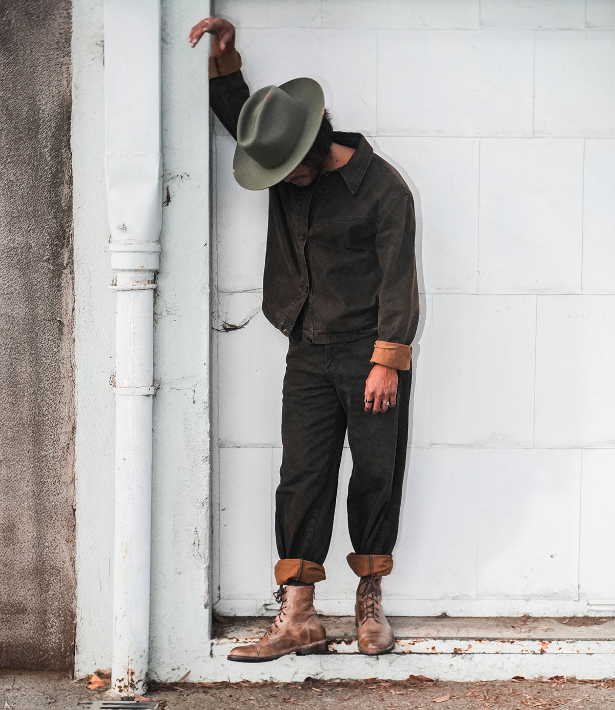 Wearing the Bed|Stü Protege boots, a person in a dark jacket, pants, and hat stands leaning against a white wall with one arm resting above their head.