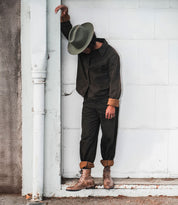 Wearing the Bed|Stü Protege boots, a person in a dark jacket, pants, and hat stands leaning against a white wall with one arm resting above their head.